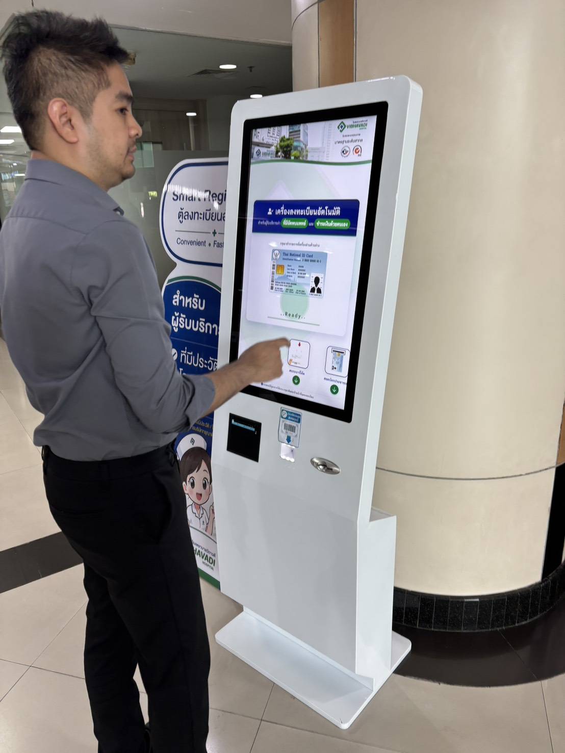 เจ้าหน้าที่โรงพยาบาลช่วยแนะนำผู้สูงอายุใช้ตู้ Kiosk หน้าจอสัมผัสขนาดใหญ่ ลดเวลารอคิว
