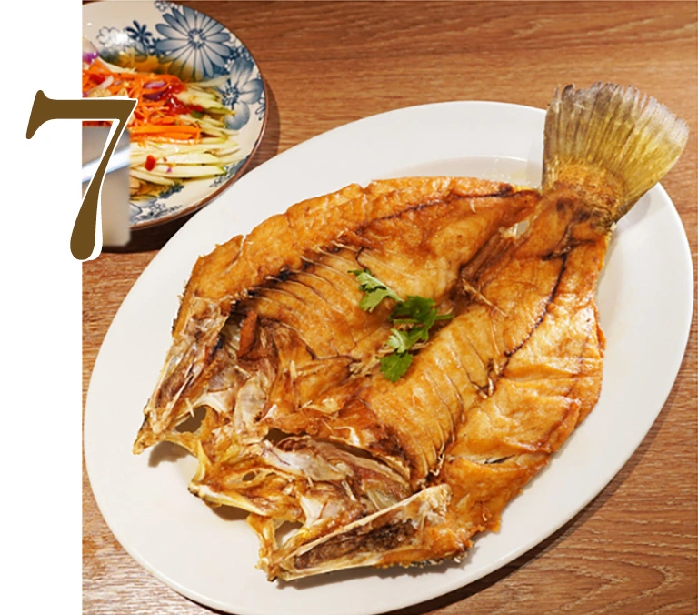 A whole golden-brown Deep Fried Sea Bass served on a white plate with a side of Spicy Mango Salad, featuring fresh mango strips and Thai herbs at Hua Seng Hong restaurant.