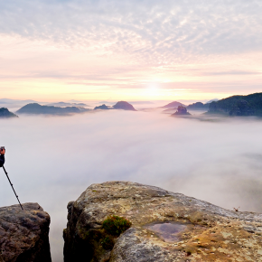 Professional nature photographer over clouds. Man takes photos with camera on tripod on rocky peak.