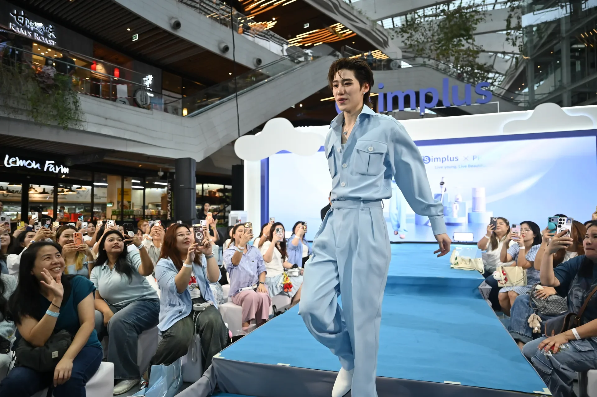 ผู้แสดงเดินบน runway สีฟ้า ผู้ชมสองฝั่ง และเวที Simplus ด้านหลัง