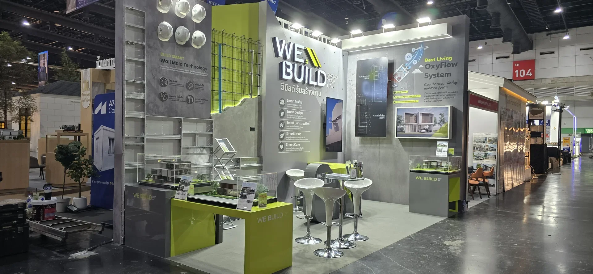 บูธ WE BUILD เสร็จแล้ว มุมเฉียงเห็นผนัง โต๊ะ โมเดลและจอภาพ
