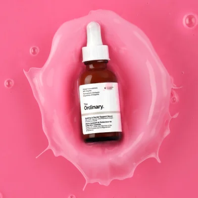 ขวดเซรั่ม The Ordinary Soothing & Barrier Support Serum ตกลงบนของเหลวสีชมพูจนเกิดเป็นรูปสแปลช