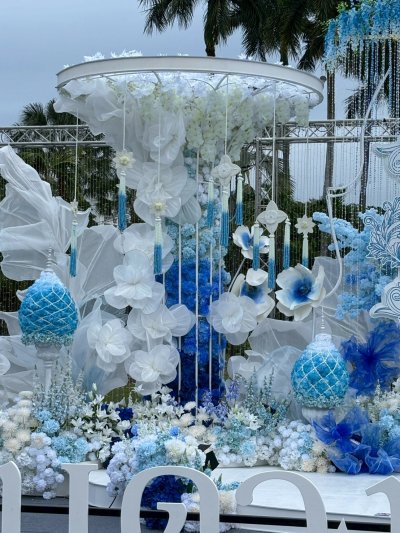 รายละเอียดงานตกแต่งซุ้มพิธีการด้วยดอกไม้โทนสีฟ้าและขาว สำหรับงานกาชาดประจำปี จัดวางตามลำดับพิธีการ